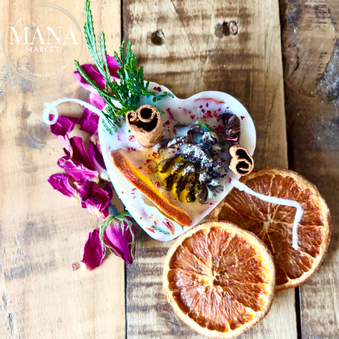 A heart-shaped fire starter made with soy wax, infused with various scents, and adorned with dried flowers, herbs, and a pine cone, displayed on a wooden surface.