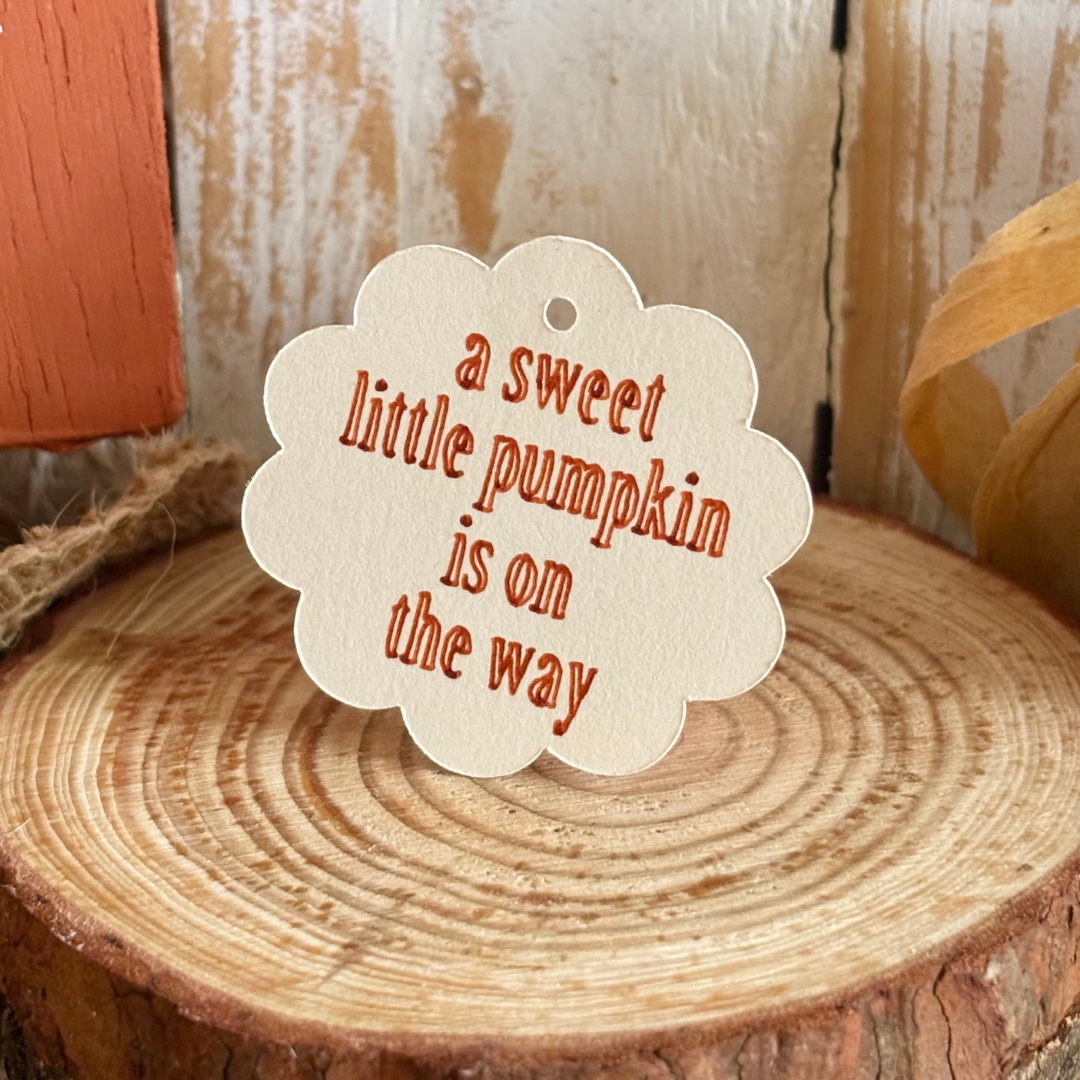 Decorative tag with 'a sweet little pumpkin is on the way' text on a wooden surface