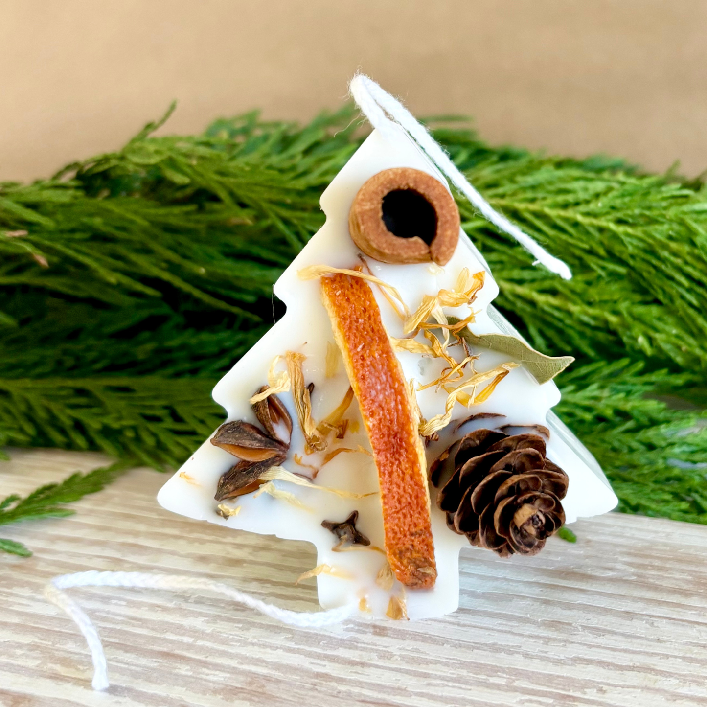 Decorative candle shaped like a Christmas tree with pine cones and orange peel on a wooden surface with greenery in the background.