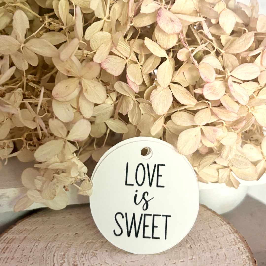 Round white tag with 'Love is Sweet' text on a background of dried flowers.