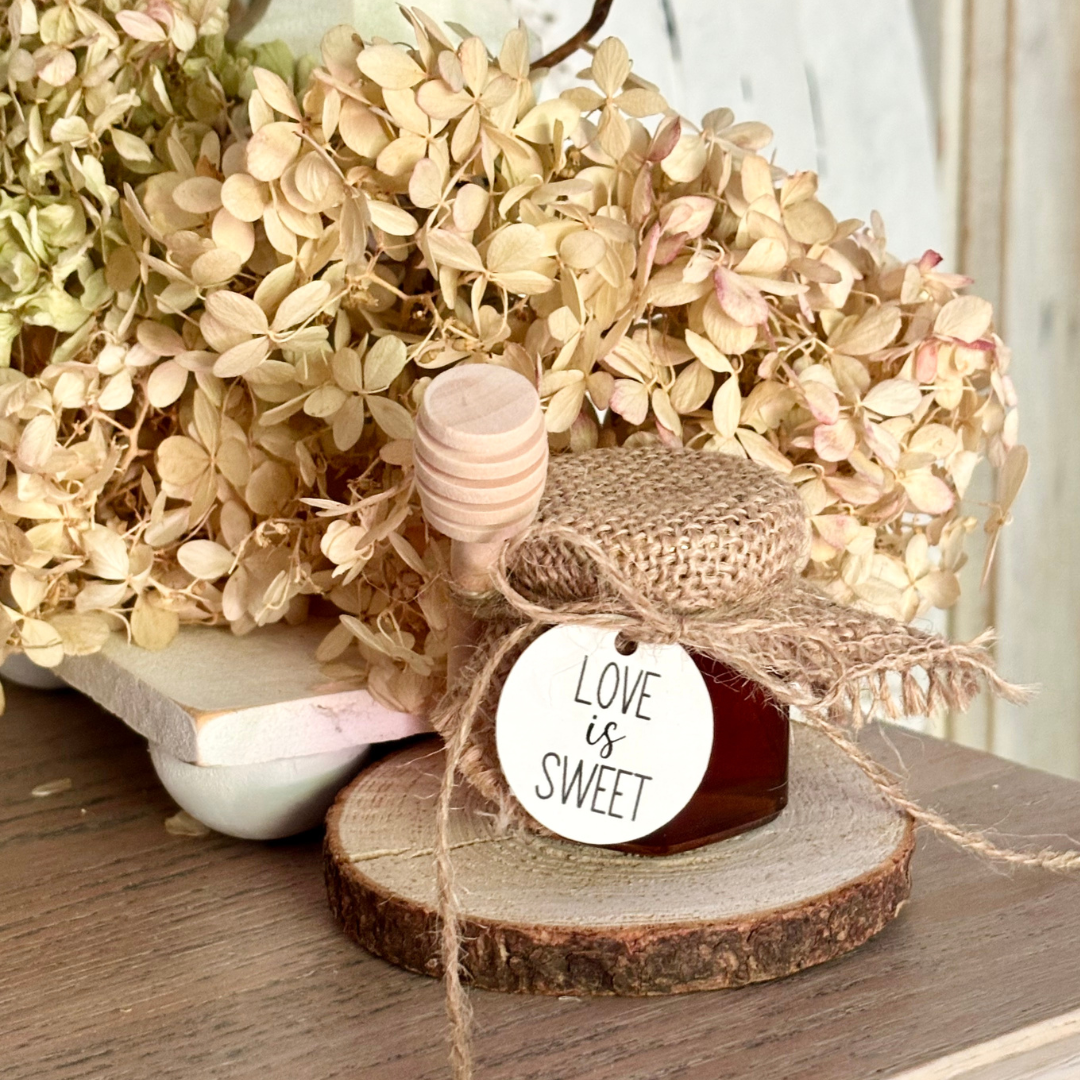 Small jar with a 'Love is Sweet' tag on a wooden slice, surrounded by flowers.