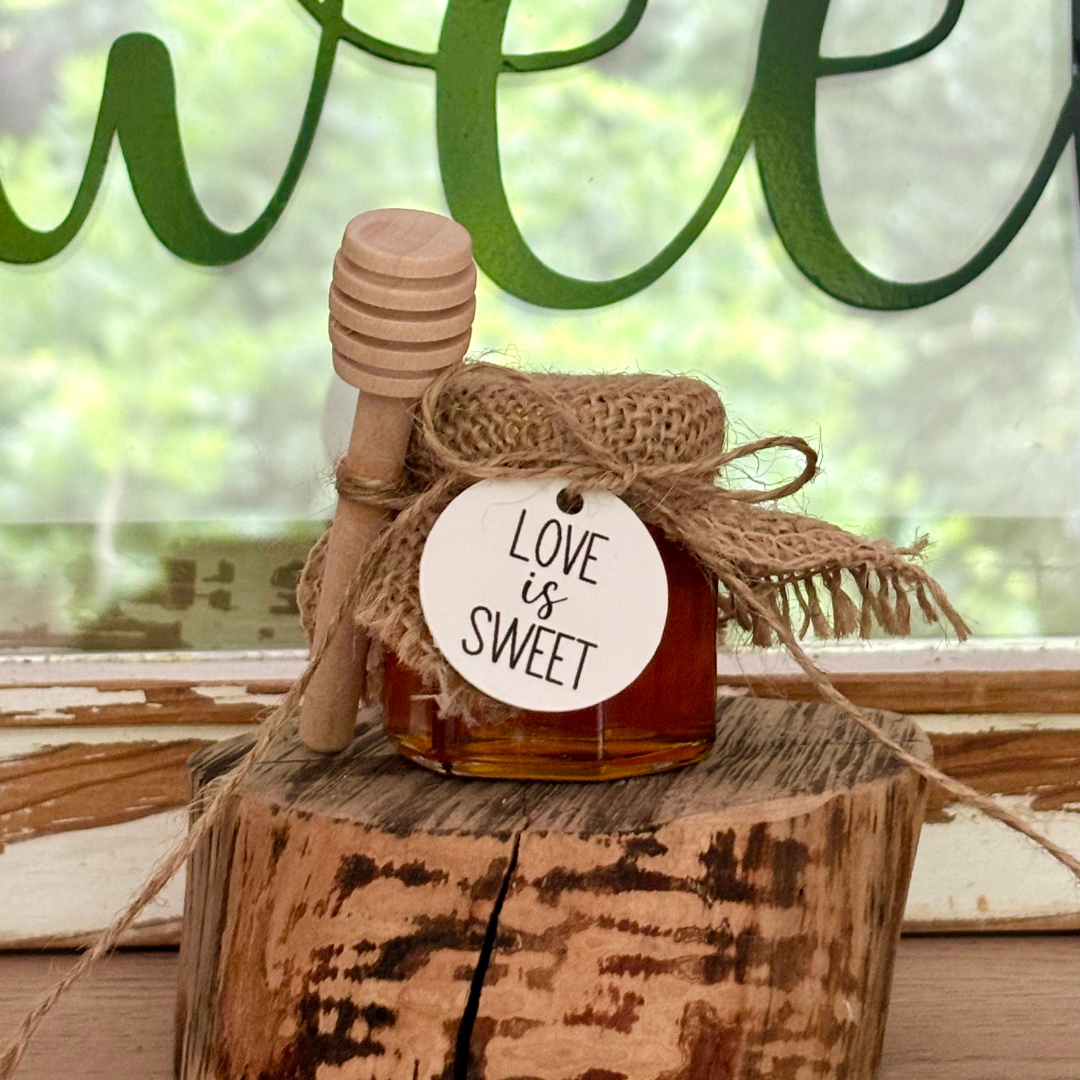 Jar of honey with a wooden dipper on a wooden block, tied with burlap and a 'Love is Sweet' tag, against a blurred green and brown background.